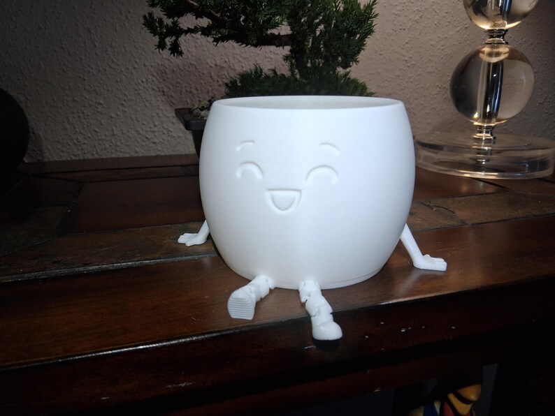 May include: A white, spherical plant pot with a smiling face design. The pot has small, attached legs and arms. The pot is made of a smooth material and is designed to hold small plants or succulents. The overall theme is whimsical and decorative.