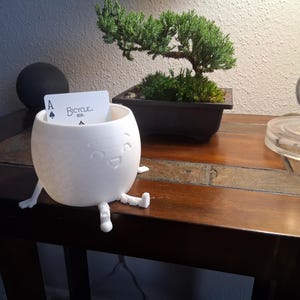 May include: A white, egg-shaped container with a smiling face and small legs. It holds a playing card with the Ace of Spades. The word "Bicycle" is visible on the card. The container is on a dark wooden table.