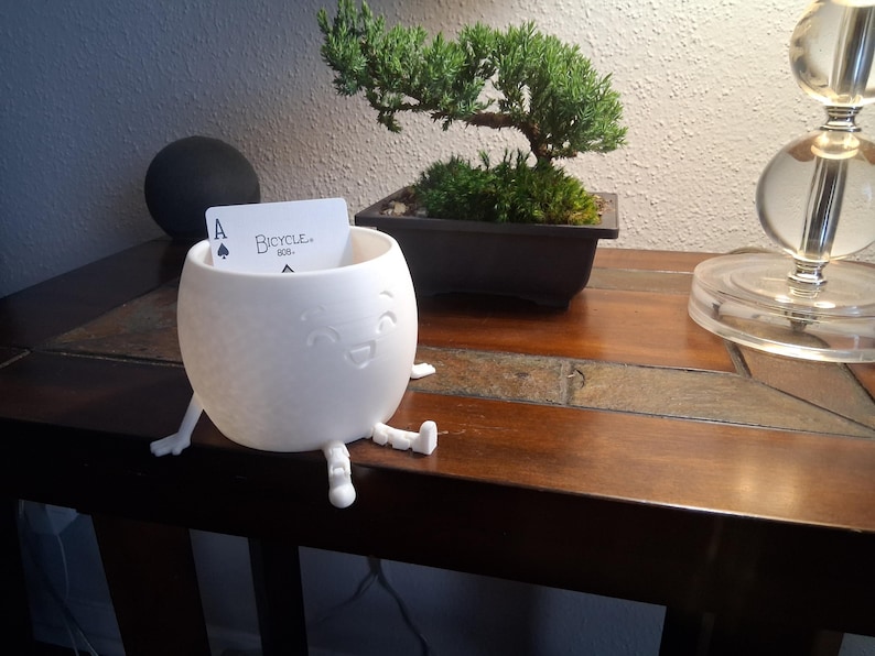 May include: A white, egg-shaped container with a smiling face and small legs. It holds a playing card with the Ace of Spades. The word "Bicycle" is visible on the card. The container is on a dark wooden table.
