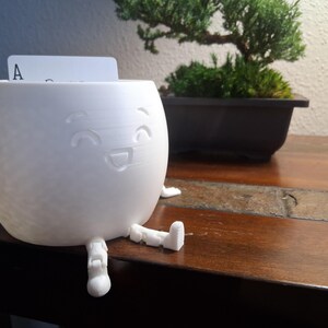 May include: A white, spherical container with a smiling face design and small legs. A playing card with the letter "A" is inserted in the top opening. The container is on a wooden surface, with a bonsai tree in the background.