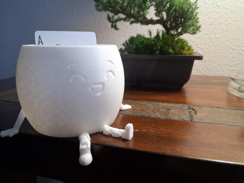 May include: A white, spherical container with a smiling face design and small legs. A playing card with the letter "A" is inserted in the top opening. The container is on a wooden surface, with a bonsai tree in the background.