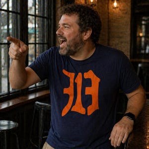 May include: A navy blue t-shirt with an orange stylized "D" logo. The person is wearing a black watch and khaki pants. The background includes a window, bar stools, and warm lighting.