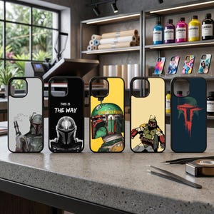May include: Five phone cases with black borders, each featuring a different design. The designs include images of a character in a helmet, the text "THIS IS THE WAY", and illustrations of a character in a green helmet and armor. The cases are displayed on a gray surface.