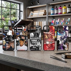 May include: Five black phone cases with various graphic designs. The cases feature images of musicians and album art. The cases are displayed on a gray countertop in a shop setting. Other cases are displayed on a shelf in the background.