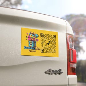 May include: A rectangular yellow magnet attached to a white vehicle tailgate. The magnet features a cartoon gift box with clothing items, the text DressupBox Apparel, and a black QR code with a small dinosaur icon in the centre of the pattern.