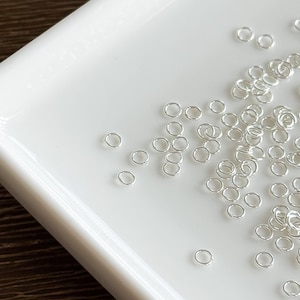 May include: A white rectangular tray filled with small, silver jump rings. The jump rings are scattered across the tray. Some are clustered together, others spaced apart. The tray has a glossy finish and rounded edges. The jump rings are likely used for jewellery making.