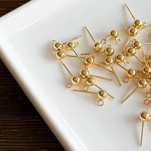 May include: A collection of small, gold-colored earring posts with ball-shaped ends and small loops. The posts are scattered on a white, rectangular tray, creating a bright and reflective display. The jewelry components are ideal for DIY earring projects.