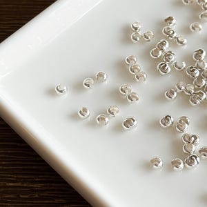 May include: Close-up of small, shiny silver crimp beads scattered on a white surface. These beads are used in jewelry making to secure clasps and other components. The beads have a smooth, rounded shape.