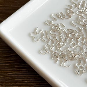 Peut inclure: Gros plan sur une collection de petits fermoirs métalliques argentés en forme de U, avec une boucle, disposés sur un plateau blanc rectangulaire. Le plateau est posé sur une surface en bois sombre.