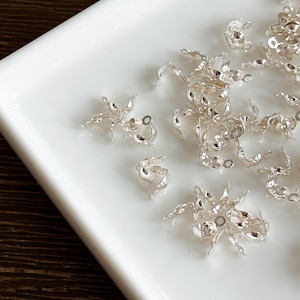 May include: A collection of small, silver-coloured metal components, likely jewellery findings, arranged on a white surface. The components feature a delicate, textured design with multiple points and small circular openings.