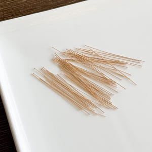 Peut inclure: Un ensemble d'épingles à tête dorées, dispersées sur une surface blanche. Les épingles ont une tige droite et fine avec une petite tête arrondie. Ces épingles sont probablement utilisées pour la fabrication de bijoux ou l'artisanat.