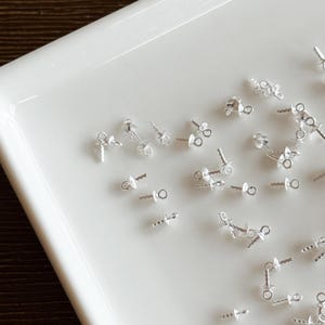 May include: A collection of small, silver-colored jewelry findings, including posts, chains, and decorative elements, are arranged on a white, rectangular tray. The findings appear to be components for earrings or other small jewelry items.