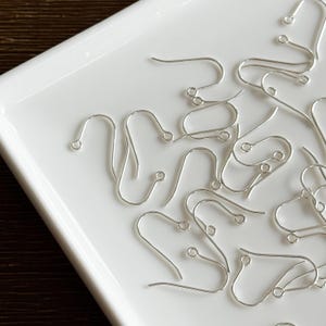 May include: A selection of silver earring hooks, arranged on a white, rectangular surface. The hooks are crafted from thin wire and feature a curved design with a small loop at the end. These hooks are for creating earrings.