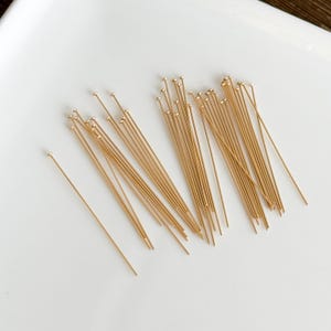 Peut inclure: Un ensemble d'épingles à tête dorées avec de petites têtes rondes. Les épingles sont dispersées sur une surface blanche, certaines regroupées et d'autres isolées. Ces épingles sont probablement utilisées pour la fabrication de bijoux ou l'artisanat.