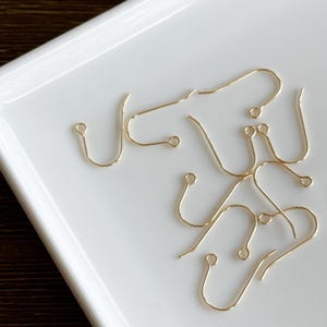 May include: A selection of gold-coloured earring hooks, arranged on a white surface. The hooks are crafted from thin wire and feature a simple, curved design with a small loop at the end.