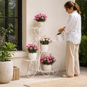 Può includere: Un portapiante in ferro battuto bianco con più livelli, ognuno con un vaso di rose rosa vibranti. Il supporto ha un design decorativo e arricciato. Una persona sta innaffiando le piante con un annaffiatoio bianco. La scena è ambientata all'aperto.