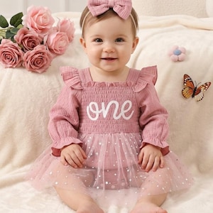 Pode incluir: Um bebê vestindo um vestido rosa com a palavra "one" em escrita branca. O vestido tem mangas compridas, uma saia de tule com estrelas e uma faixa de cabelo combinando com um laço. Uma borboleta e rosas cor-de-rosa estão no fundo.
