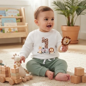 Può includere: Una felpa bianca a maniche lunghe con una grafica del numero uno con motivo a giraffa e una collezione di animali da safari illustrati, tra cui una giraffa, una zebra, un elefante e un leone, abbinata a pantaloni color verde salvia.