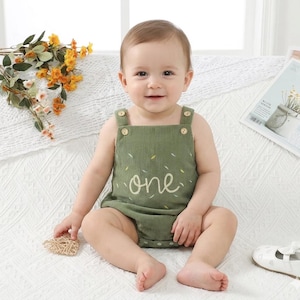 May include: A baby in a green corduroy romper with the word "one" embroidered on the front. The romper features wooden button details on the straps. The baby is sitting on a white surface.