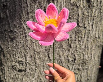 May include: A handcrafted pink lotus flower made from fuzzy pipe cleaners with a bright yellow center. The flower features multiple layered petals in a vibrant shade of pink, radiating outward from a textured yellow core, attached to a thin green stem.