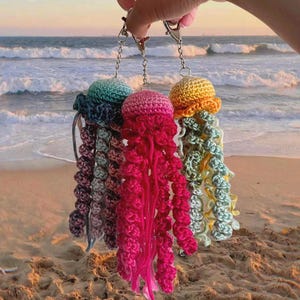 May include: Three handmade crochet jellyfish keychains featuring silver-toned metal lobster clasps and chains. The jellyfish are crafted in vibrant yarn colors including teal, magenta, and yellow, with long, spiraling, and dangling tentacles.