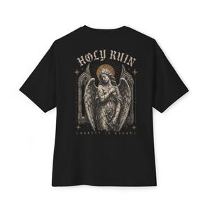 Pode incluir: T-shirt preta com um design gráfico bege e dourado de um anjo com asas, um halo e as palavras "HOLY RUIN" na parte superior. O texto "+ BEAUTY IN DECAY +" está abaixo. O design tem uma estética vintage.