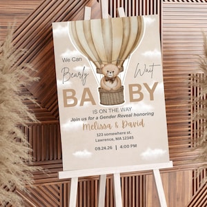 May include: A baby shower announcement on a white easel. The design features a hot air balloon with a teddy bear in the basket. Text reads "We Can Bearly Wait BABY" and includes event details for Melissa & David.