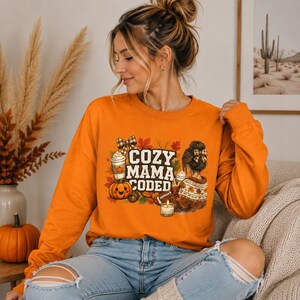 May include: Orange long-sleeve sweatshirt with the text "COZY MAMA CODED" in white. The graphic includes a pumpkin, coffee cups, fall leaves, and a woman with a bow in her hair. The sweatshirt is a fall-themed item.