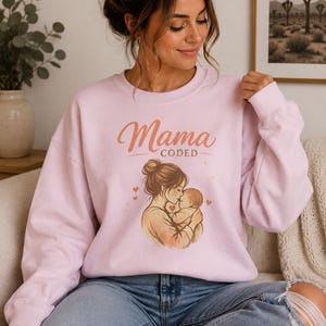 May include: A light pink sweatshirt with the word "Mama" in cursive and "CODED" in a sans-serif font. The graphic depicts a mother kissing a baby, surrounded by small hearts. The sweatshirt is worn by a person wearing blue jeans.