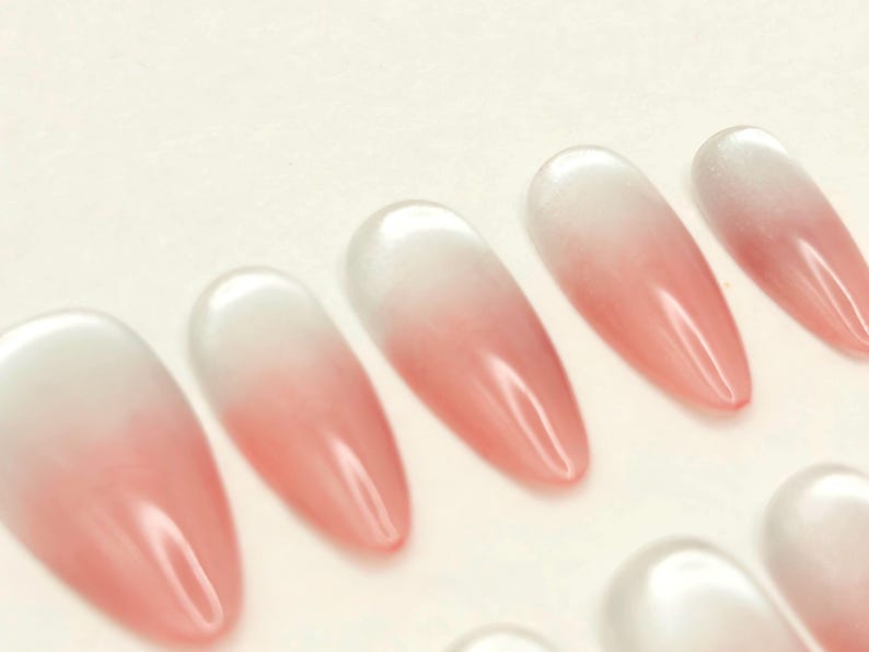 May include: A collection of almond-shaped artificial nails. Each nail features a gradient design, transitioning from a white base to a soft pink hue. The nails have a glossy finish, reflecting light.