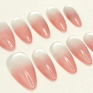 May include: A set of ten almond-shaped false nails with a gradient design. The nails transition from a white base to a soft pink hue, creating a subtle ombre effect. The nails are glossy and appear to be made of a gel-like material.