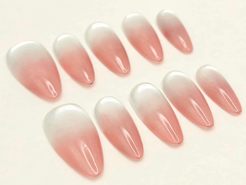 May include: A set of ten almond-shaped false nails with a gradient design. The nails transition from a white base to a soft pink hue, creating a subtle ombre effect. The nails are glossy and appear to be made of a gel-like material.