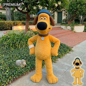 以下が含まれることがあります： 黄色い体、茶色の耳、青い帽子が特徴の、犬のキャラクターの全身コスチューム。犬は大きな黒い鼻、銀色のタグが付いた茶色の首輪、白いリストバンドをしています。コスチュームはレンガの歩道に立っています。
