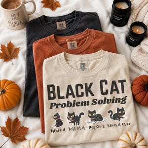 Może przedstawiać: Trzy koszulki z okrągłym dekoltem w kolorze czarnym, rdzawopomarańczowym i kremowym. Kremowa koszulka zawiera napis "BLACK CAT Problem Solving" oraz ilustracje kota wykonującego czynności "Ignore it", "Yell at it", "Nap on it" i "Knock it over".