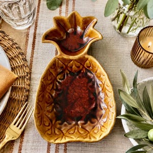 Puede incluir: Un plato de servicio de cerámica con forma de piña que presenta un exterior amarillo mostaza brillante con un patrón texturizado de escamas y un centro esmaltado en rojo oscuro. El plato tiene una parte superior en forma de hoja.