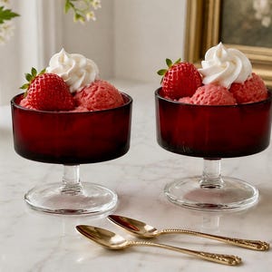 May include: Two deep red glass dessert bowls on clear stems, each filled with pink strawberry sorbet, topped with a fresh whole strawberry and a swirl of white whipped cream, accompanied by two gold-toned metal spoons with ornate handles.