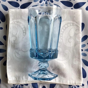 May include: A light blue glass goblet featuring a faceted, paneled bowl and a hexagonal stem base. The drinking vessel has a clear, translucent appearance with vertical panels that extend from the rim down to the base, creating a geometric design.