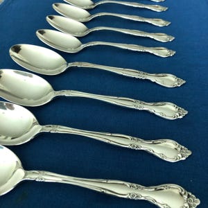 May include: A set of eight silver-toned metal spoons arranged in a diagonal row. Each spoon features a polished oval bowl and a handle decorated with embossed floral detailing at the base and along the stem, reflecting light on the surface.