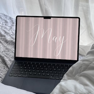 Puede incluir: Un portátil gris oscuro muestra la palabra "May" en escritura blanca. La pantalla tiene un fondo de rayas verticales rosas y grises. El portátil está sobre una cama con sábanas con estampado floral.