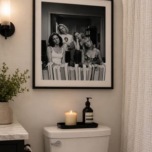 May include: Monochrome framed print featuring five women seated behind books. A lit candle, soap dispenser, and a black tray are on a white toilet. A wall light and a plant in a vase are also visible in the bathroom.
