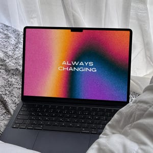 Puede incluir: Un portátil negro que muestra el texto "ALWAYS CHANGING" sobre un fondo degradado colorido de rosa, naranja, azul y morado. El portátil está abierto sobre una cama con una sábana estampada floral y un edredón blanco.