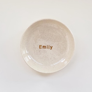 May include: A round, off-white ceramic trinket dish with a speckled texture throughout the surface. The name Emily is printed in the center of the dish in a simple, dark brown sans-serif font. The dish has a smooth, glossy finish and raised edges.