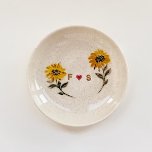 May include: A round, speckled off-white ceramic trinket dish featuring two hand-painted yellow sunflowers with green stems and leaves. Between the flowers, the letters F and S are printed in a dark font, separated by a small red heart.