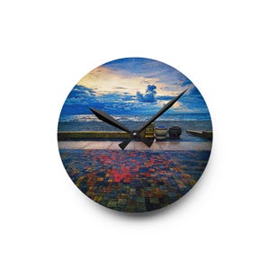 May include: A round wall clock featuring a printed image of a seaside promenade at dusk. The foreground shows a multi-coloured stone-tiled walkway leading to a low wall, the ocean, and a cloudy sky. Two black clock hands are centred on the face.