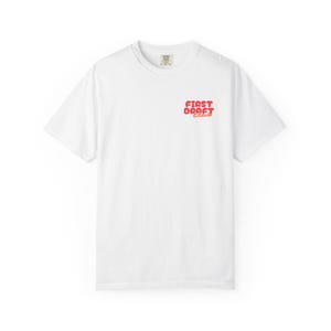 Puede incluir: Una camiseta blanca de manga corta con el texto "FIRST DRAFT CLUB" impreso en una fuente roja, negrita y redondeada en la parte superior izquierda del pecho. La prenda tiene un cuello redondo clásico y un corte estándar de diseño limpio.