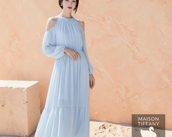 May include: A light blue, long-sleeve dress with cold shoulder cutouts. The dress has a high neckline and a flowing skirt. The image includes the text "MAISON TIFFANY STUDIO".