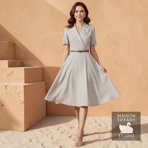 May include: A light gray dress with short sleeves, a V-neck, and a brown belt. The dress has a flared skirt and a tailored bodice. The image includes the "MAISON TIFFANY STUDIO" logo.