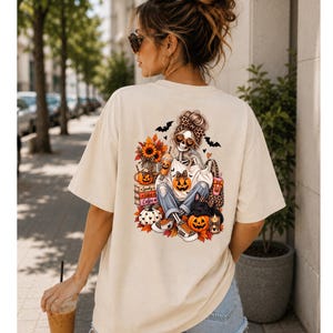 Könnte beinhalten: Ein beiges Kurzarm-T-Shirt mit einer farbenfrohen Grafik auf der Rückseite. Das Design zeigt ein Skelett mit Dutt, das eine Sonnenbrille und ein Leoparden-Stirnband trägt, umgeben von Kürbissen, Sonnenblumen, Büchern und Fledermäusen.