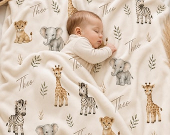 May include: A cream-colored fleece blanket featuring a repeating pattern of watercolor-style illustrations of giraffes, elephants, zebras, and lion cubs, interspersed with green leafy sprigs and the name Theo written in a cursive script.
