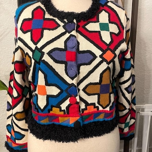 May include: A long-sleeve knit cardigan featuring a cream base with a bold geometric pattern in red, blue, purple, orange, and green. The garment has a black fuzzy trim along the neckline, cuffs, and hem, and includes three round, textured buttons.
